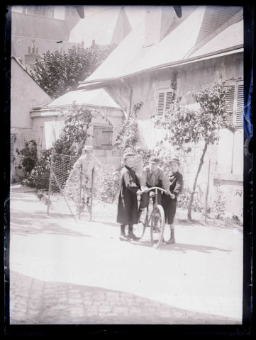 Jeunes garçons à bicyclette et sur des échasses devant une habitation.