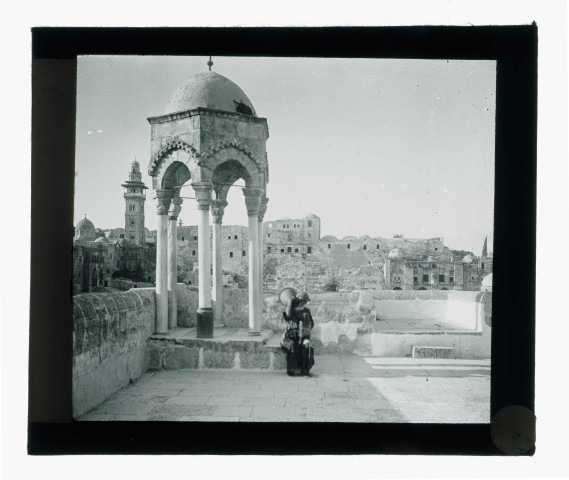 Porteuse d'eau sur l'esplanade d'une mosquée avec mihrab et minaret en arrière plan.