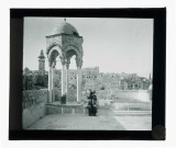 Porteuse d'eau sur l'esplanade d'une mosquée avec mihrab et minaret en arrière plan.