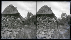 Réserve de bois le long du pignon d'une chaumière normande et clocher au loin.