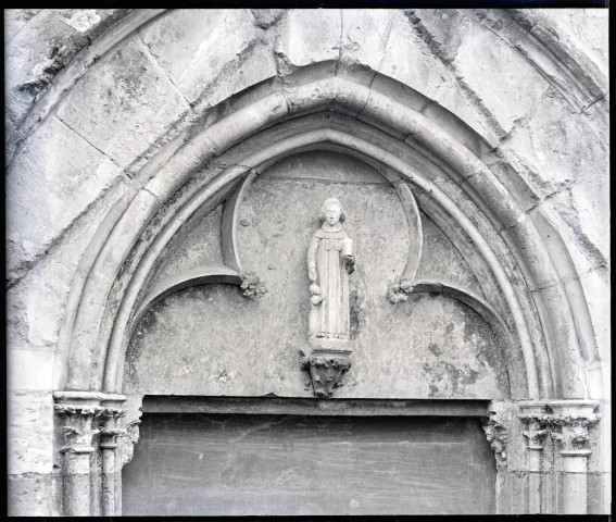 Statue en calcaire du XIVe siècle, sur la façade Nord de l'église Notre-Dame d'Écouis (Eure), sous l'arc trilobé du tympan surmontant la porte, et représentant probablement Saint-Côme tenant une boîte et accompagné d'un enfant