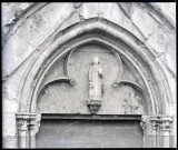 Statue en calcaire du XIVe siècle, sur la façade Nord de l'église Notre-Dame d'Écouis (Eure), sous l'arc trilobé du tympan surmontant la porte, et représentant probablement Saint-Côme tenant une boîte et accompagné d'un enfant