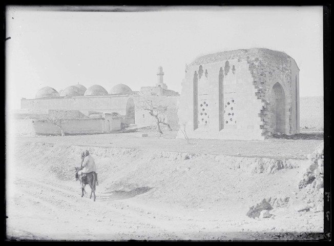 Deux hommes à dos d'âne sur un chemin de campagne avec à l'arrière-plan un minaret et des mausolées sous coupole.
