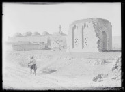 Deux hommes à dos d'âne sur un chemin de campagne avec à l'arrière-plan un minaret et des mausolées sous coupole.