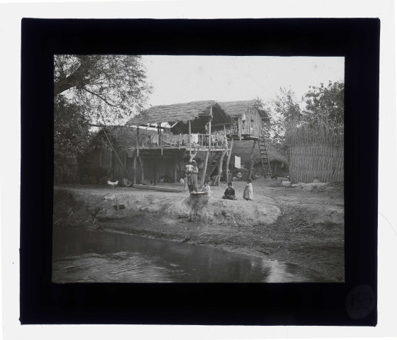 Enfants dans une cour de ferme au bord d'une rivière avec les maisons sur pilotis.