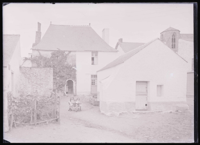 Jeunes enfants dans une brouette devant une habitation de bourgade.