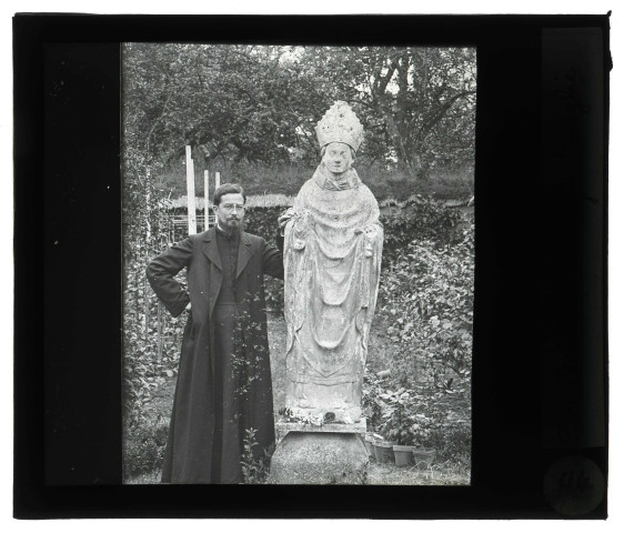 Jardin du presbytère de Saint-Aubin-du-Thenney (Eure), un prêtre (M. Machal) est appuyé contre la statue du Saint-Évêque.