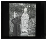 Jardin du presbytère de Saint-Aubin-du-Thenney (Eure), un prêtre (M. Machal) est appuyé contre la statue du Saint-Évêque.