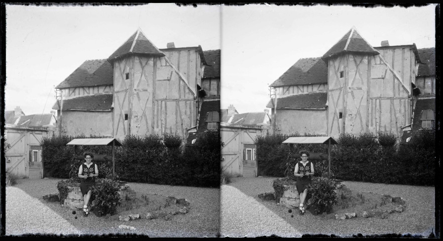 Jeune fille tricotant assise sur la margelle d'un puits devant une maison à pans de bois avec une tourelle.
