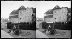 Jeune fille tricotant assise sur la margelle d'un puits devant une maison à pans de bois avec une tourelle.