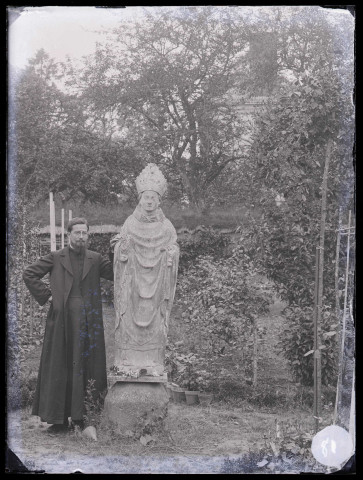 Jardin du presbytère de Saint-Aubin-du-Thenney (Eure), un prêtre (M. Machal) est appuyé contre la statue du Saint-Évêque.