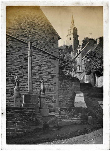 Petit calvaire breton à l'angle d'une ruelle avec le clocher de l'église au loin.