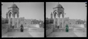 Porteuse d'eau sur l'esplanade d'une mosquée avec mihrab et minaret en arrière plan.