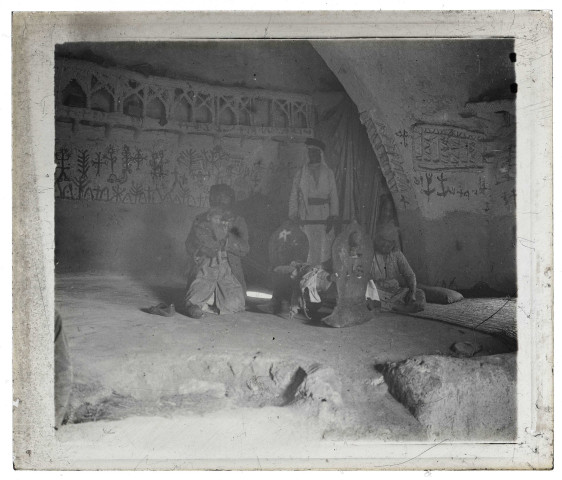 Famille de villageois autour d'un berceau dans une habitation traditionnelle au décor rupestre.