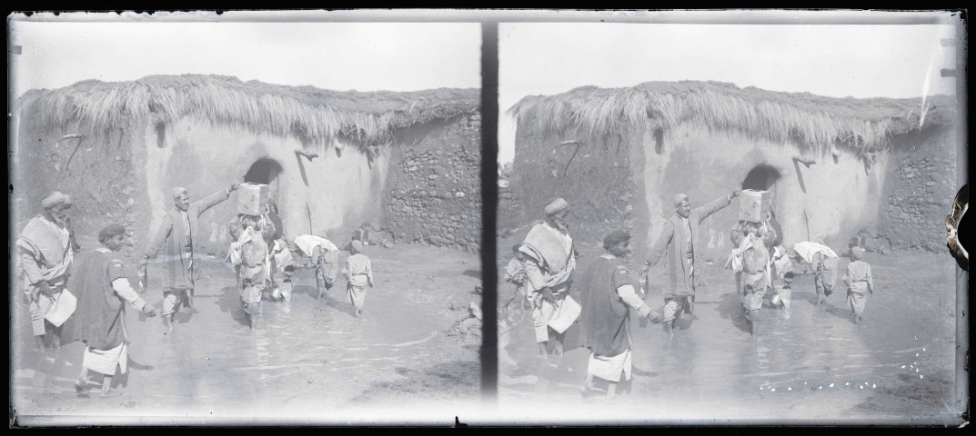 Hommes et enfants à un point d'eau devant une habitation en torchis.