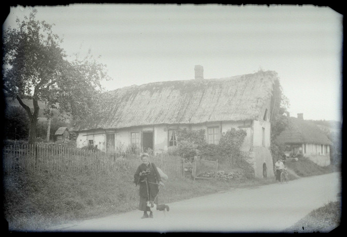 Femme et son chien sur un chemin devant une chaumière normande.