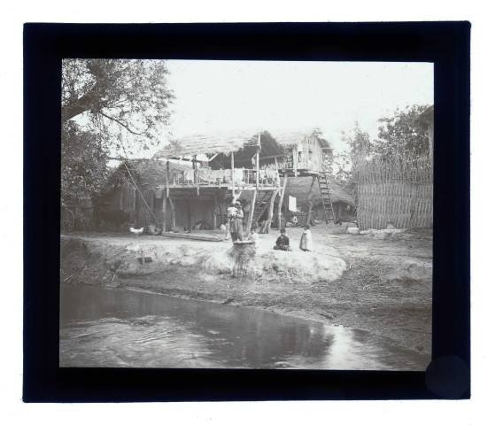 Enfants dans une cour de ferme au bord d'une rivière avec les maisons sur pilotis.