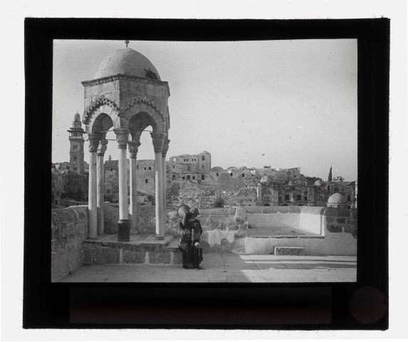 Porteuse d'eau sur l'esplanade d'une mosquée avec mihrab et minaret en arrière plan.