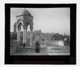 Porteuse d'eau sur l'esplanade d'une mosquée avec mihrab et minaret en arrière plan.